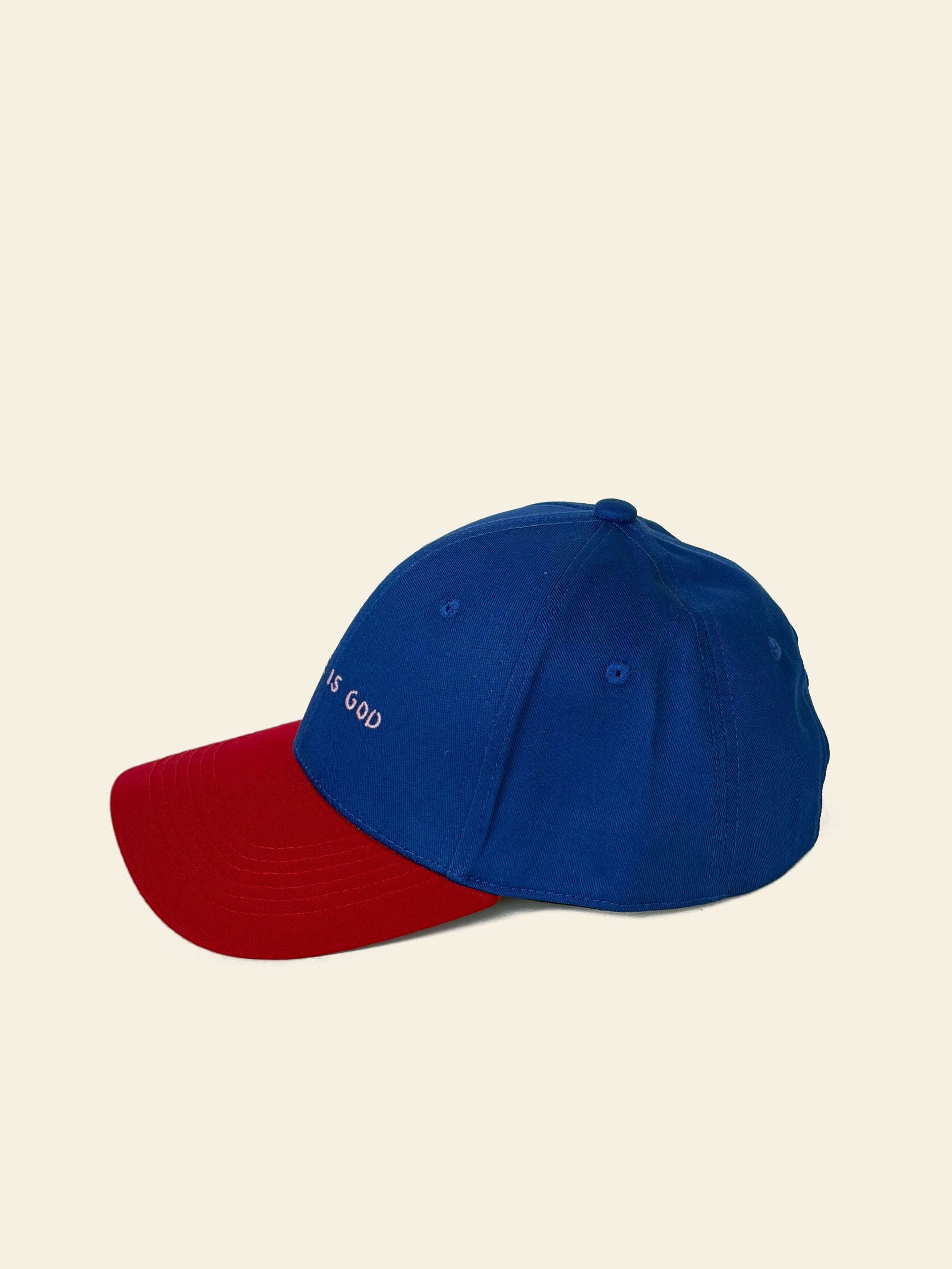 The David Hockney Space Is God Cap by David Hockney is a blue two-tone cotton hat with a red brim, featuring small white “SPACE IS GOD” embroidery on the front, photographed against a plain beige background.