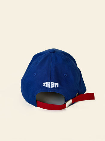 A back view of the David Hockney Space Is God Cap by David Hockney, a blue two-tone cotton cap featuring a red adjustable strap and “SPACE IS GOD” embroidered in white above the opening.