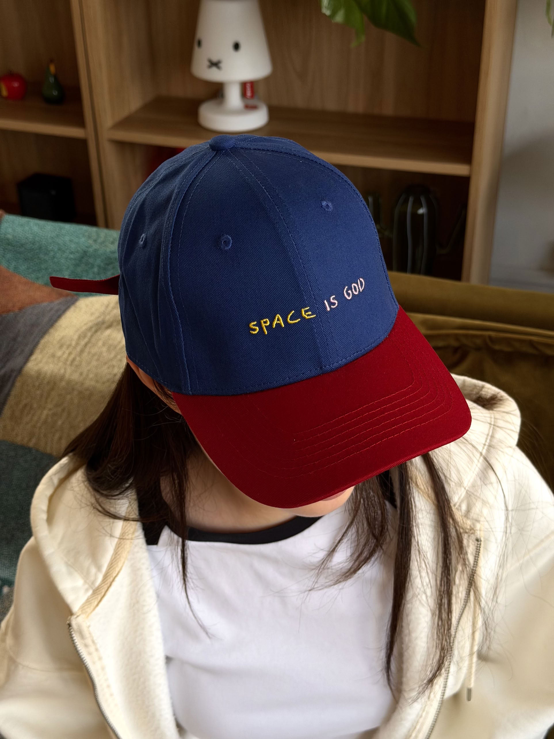 A person sits indoors on a couch, facing downward, wearing the David Hockney Space Is God Cap by David Hockney—a blue and red two-tone cotton cap featuring yellow “SPACE IS GOD” embroidered text. Shelves and a white bunny lamp appear in the background, evoking an atmosphere in line with the iconic David Hockney cap aesthetic.