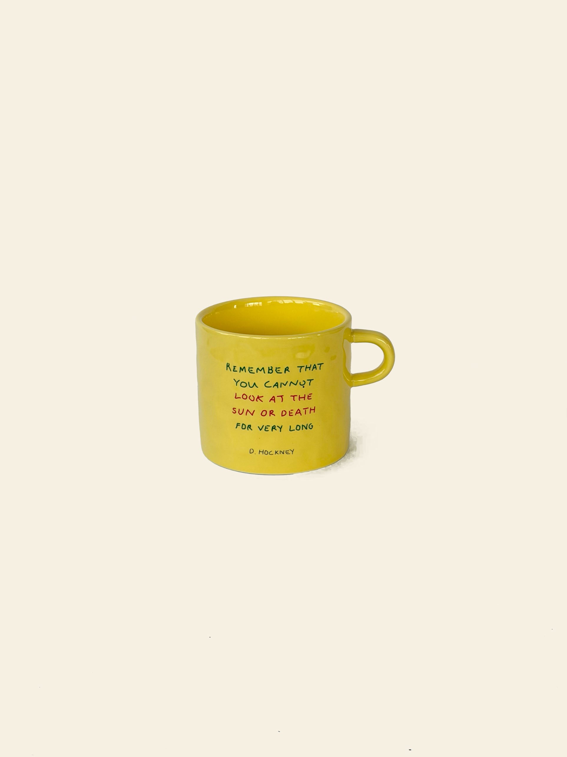 A yellow ceramic David Hockney Bigger & Closer Mug with a handle, displayed on a beige background. This mug by David Hockney features the quote: “Remember that you cannot look at the sun or death for very long.”.