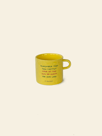 A yellow ceramic David Hockney Bigger & Closer Mug with a handle, displayed on a beige background. This mug by David Hockney features the quote: “Remember that you cannot look at the sun or death for very long.”.