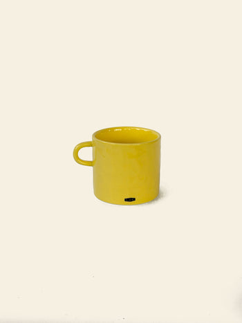 A bright yellow ceramic mug with a handle sits on a plain, light background. The David Hockney Bigger & Closer Mug by David Hockney features a subtle shine and a small black logo near its base, echoing the iconic style of Hockney’s work.