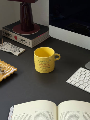 A David Hockney Bigger & Closer Mug by David Hockney, featuring yellow ceramic with writing, sits on a dark desk near a keyboard, an open book, and a stack of books—including one titled PROJECT JAPAN. A tray holds jewelry and a tea bag, and a lamp is also visible.