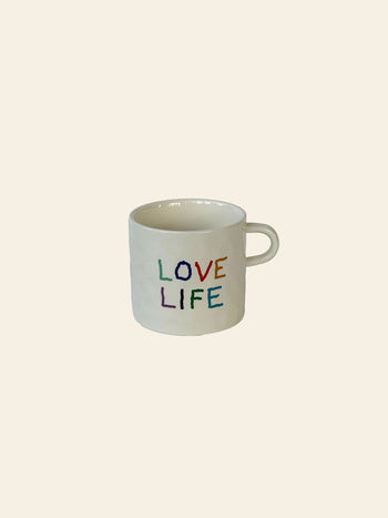 The David Hockney Love Life Mug by David Hockney is a white ceramic mug featuring “LOVE LIFE” written in colorful, hand-painted letters on the side, set against a plain, light beige background.