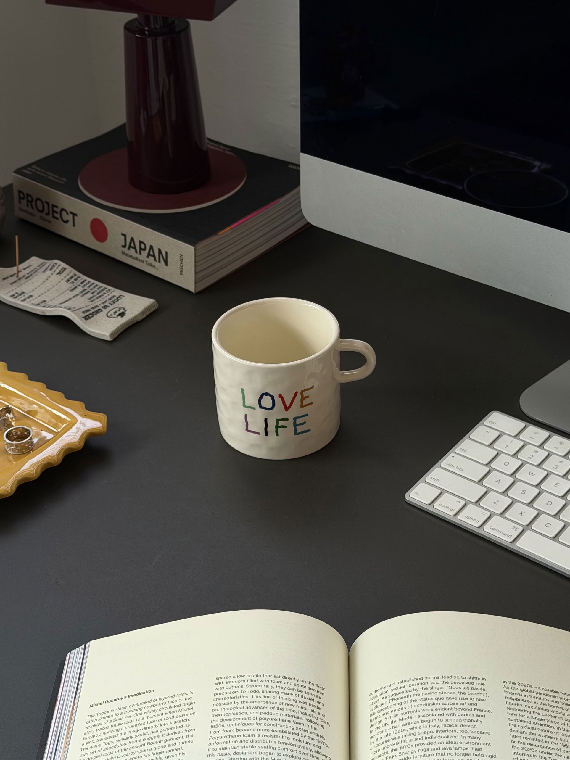 A David Hockney Love Love Mug sits on a dark desk near a keyboard, computer, open book, lamp, a tray with jewelry, a Japanese-themed book, and a small paper item.