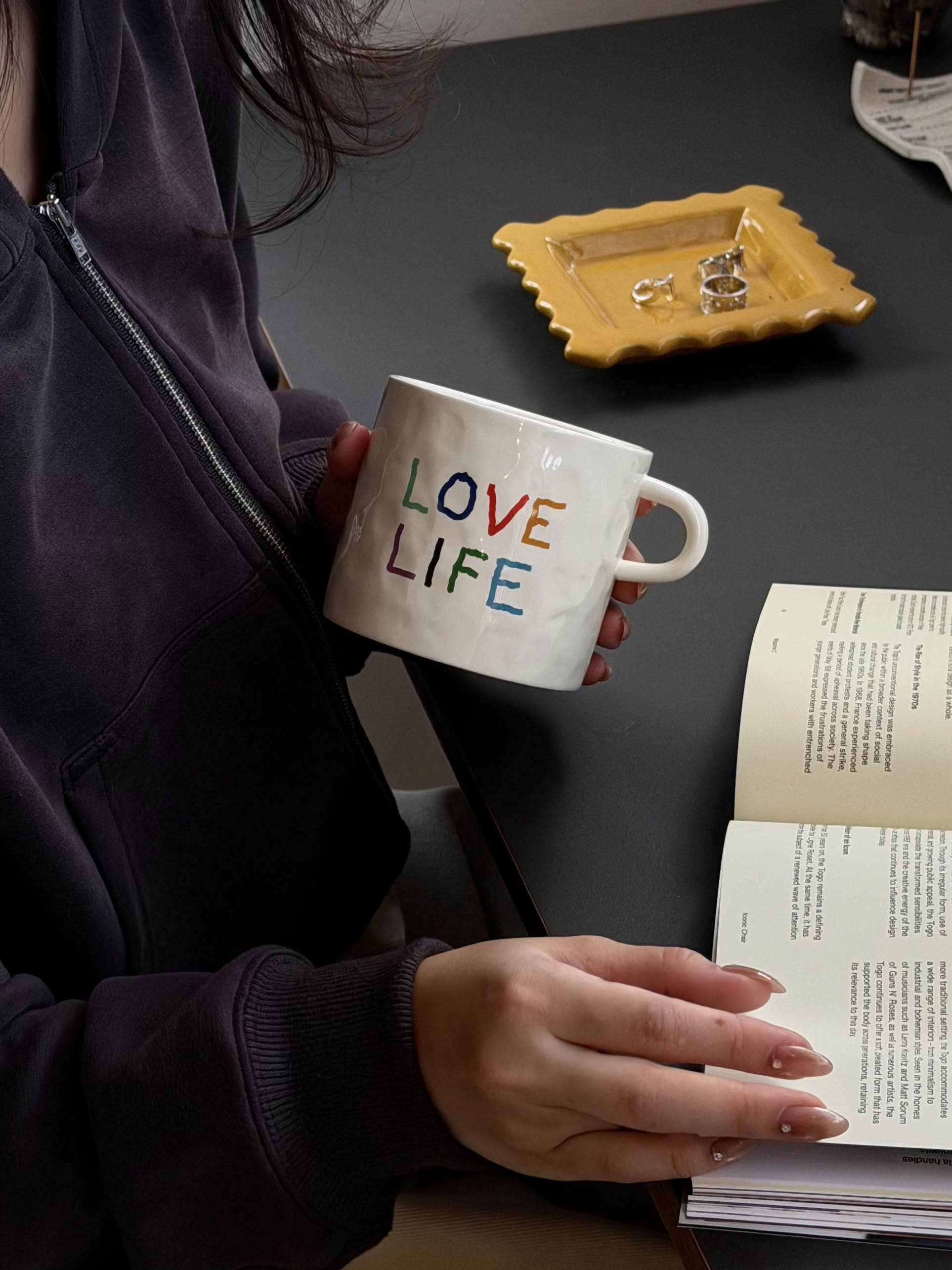 Clad in a dark hoodie, a person reads a book at a table set with a yellow dish and rings, savoring the charm of their David Hockney Love Love Mug by David Hockney.