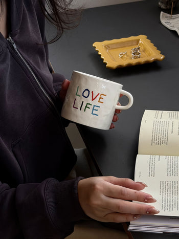 Clad in a dark hoodie, a person reads a book at a table set with a yellow dish and rings, savoring the charm of their David Hockney Love Love Mug by David Hockney.