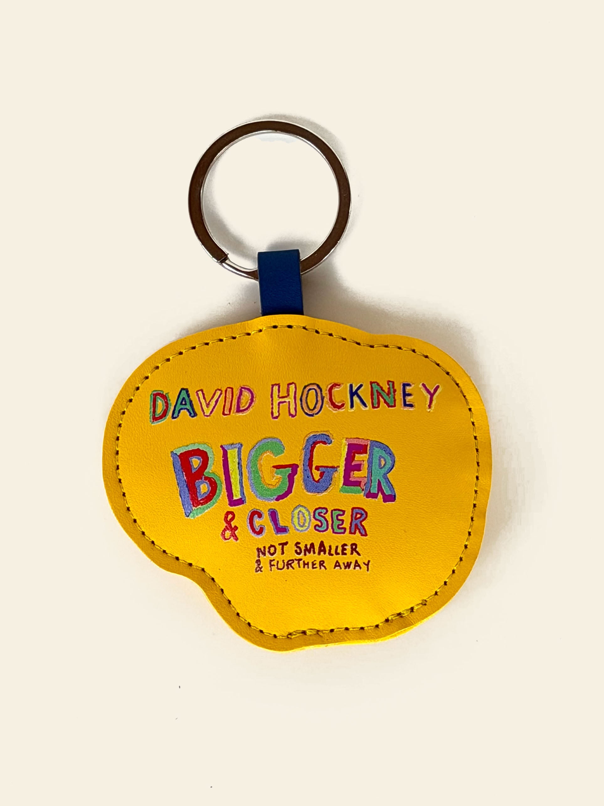 The David Hockney Bigger & Closer Keychain by David Hockney features an irregular yellow shape, colorful “Bigger & Closer (not smaller & further away)” text, and a silver metal keyring—making it a playful accessory for art lovers.