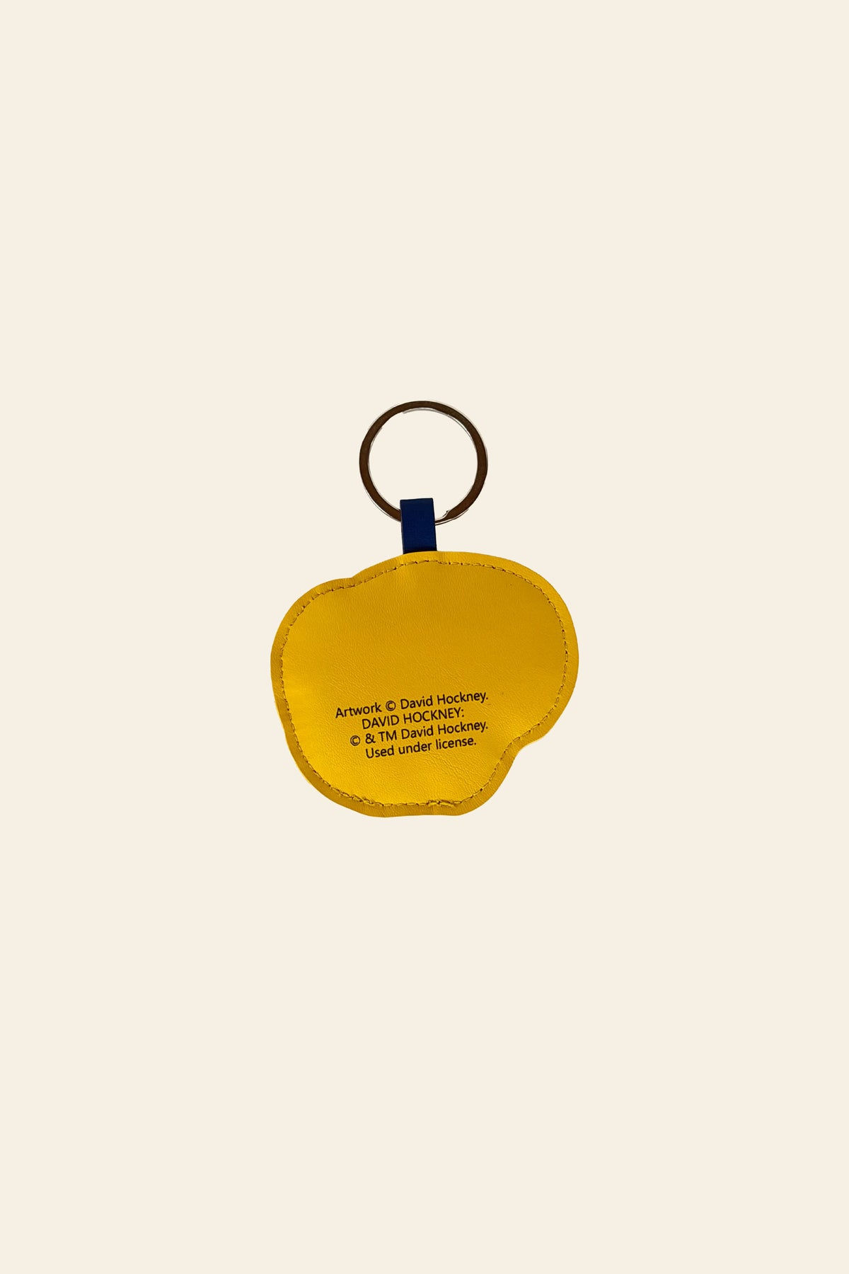 A yellow, apple-shaped David Hockney Bigger & Closer Keychain with printed text is attached to a brown metal ring and a small blue fabric loop, and is displayed on a plain beige background. This unique keychain is an exhibition souvenir from the David Hockney collection.