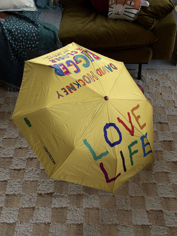 A bright yellow David Hockney Bigger & Closer Umbrella rests on a checkered rug, featuring the words “LOVE LIFE” in colorful letters and mirrored text of “DAVID HOCKNEY” and “BIGGER & CLOSER,” capturing the playful essence of the David Hockney brand. In the background, a couch with a cushion and blanket adds cozy comfort to the scene.