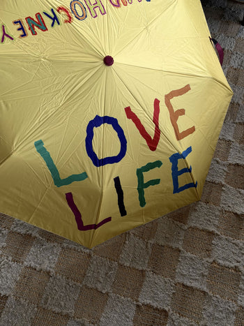 A David Hockney Bigger & Closer Umbrella by David Hockney, featuring “LOVE LIFE” painted in colorful, uneven letters on a yellow canopy, lies open on a checkered rug and textured carpet.