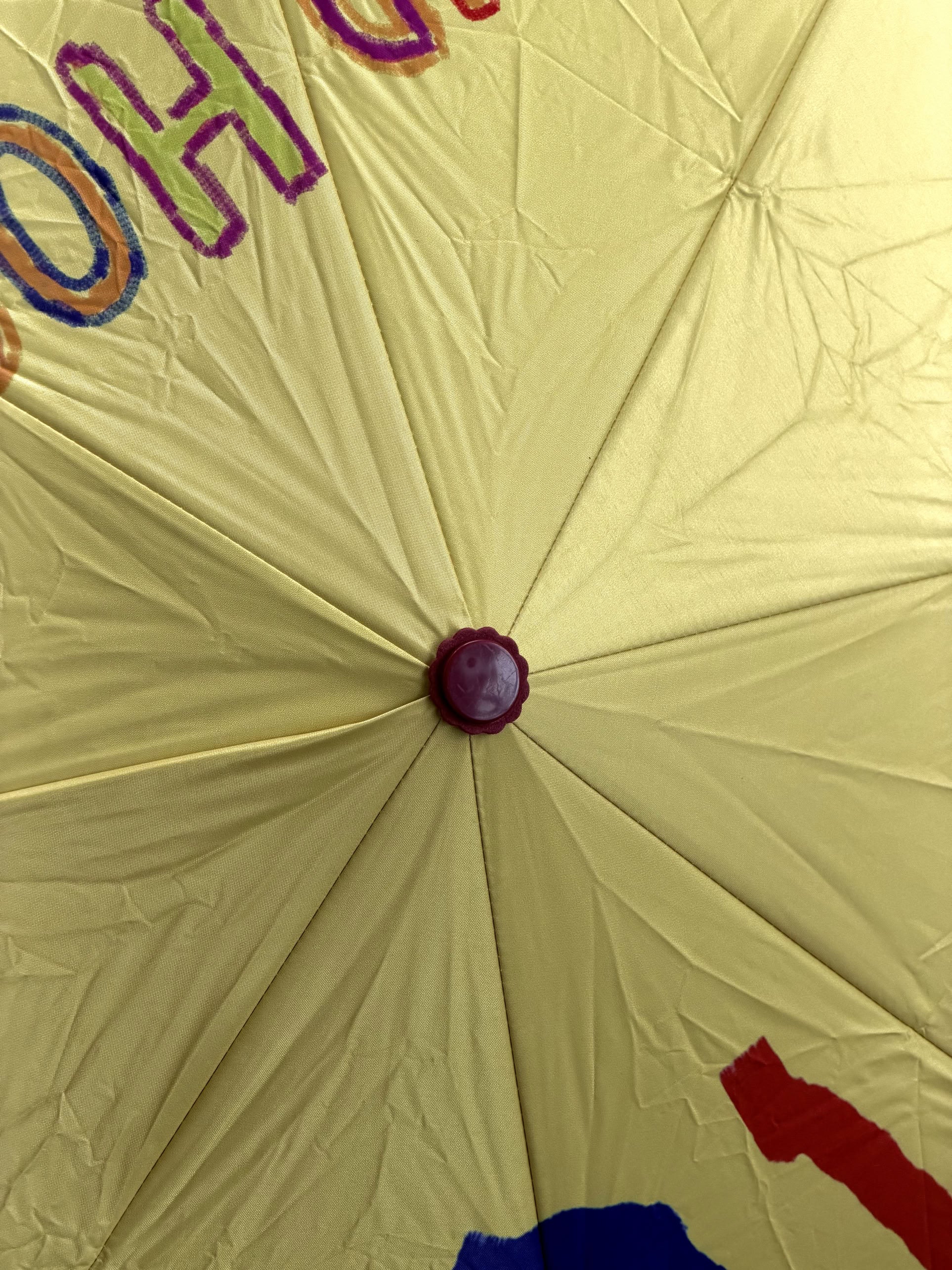 Close-up of the center of a yellow David Hockney Bigger & Closer Umbrella, with red, purple, and blue painted designs partially visible near the edges. This umbrella by David Hockney showcases vibrant artistry reminiscent of the iconic designer’s style.