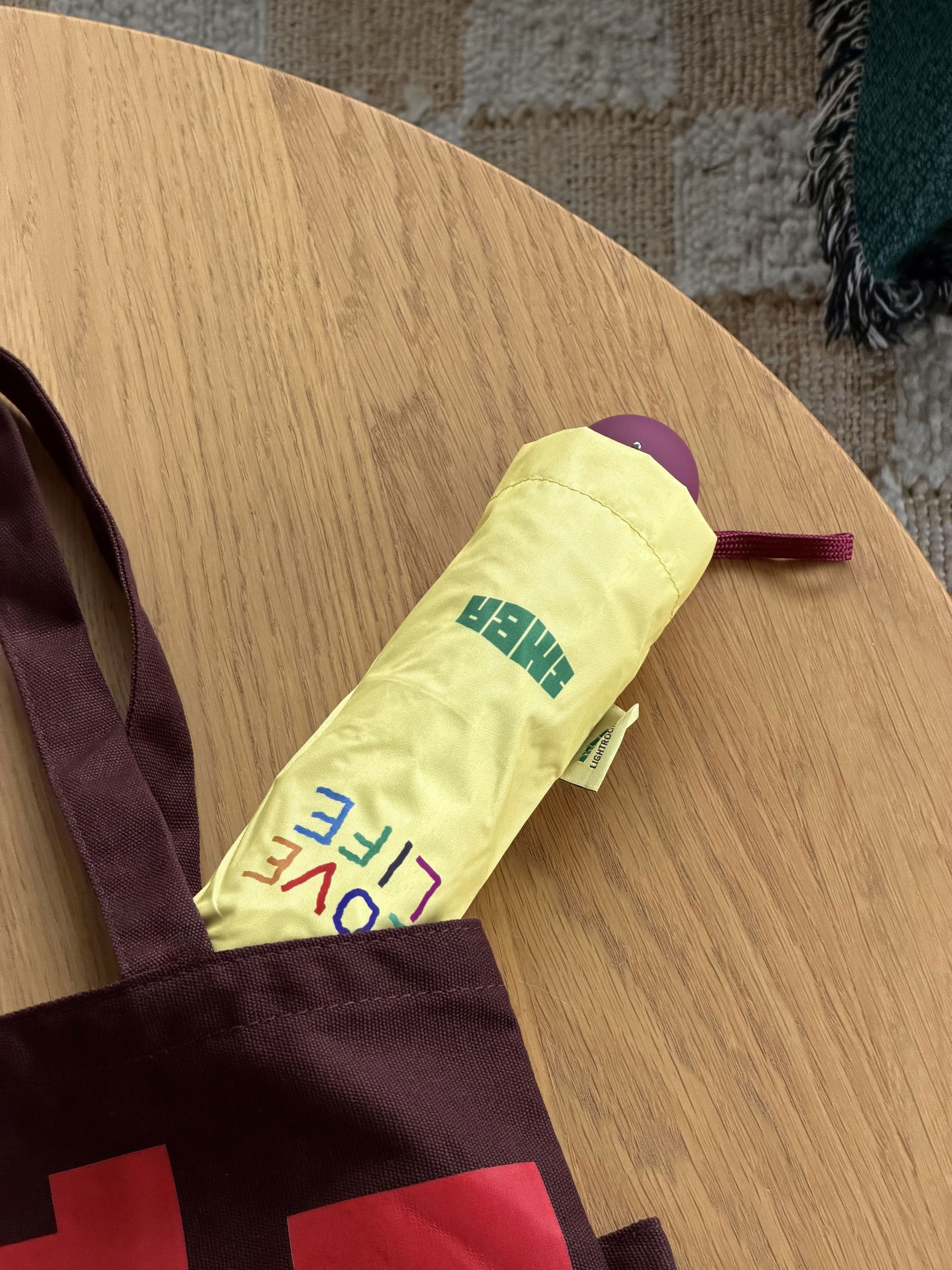 A yellow David Hockney Bigger & Closer Umbrella, featuring the words TIGER and LOVE LIFE, is partially sticking out of a maroon tote bag on a light wooden table. A rug is visible in the background.