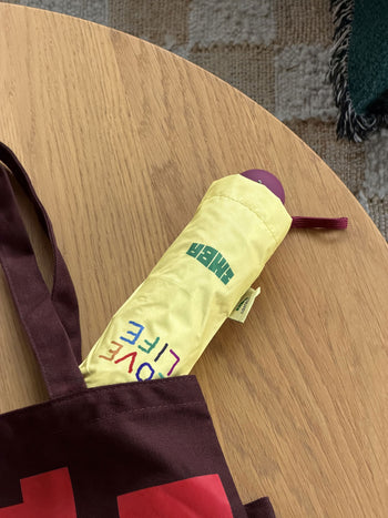 A yellow David Hockney Bigger & Closer Umbrella, featuring the words TIGER and LOVE LIFE, is partially sticking out of a maroon tote bag on a light wooden table. A rug is visible in the background.