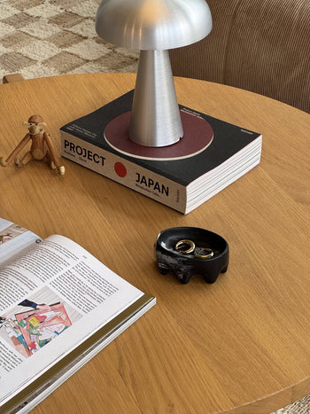 The Dinosaur Designs Crown Dish by Dinosaur Designs is a small, round, black ceramic piece with a glossy finish and four drippy legs, centered on a light beige background, echoing the brands signature sculptural style.