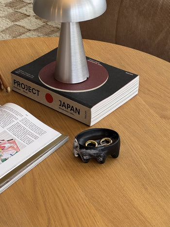 A round wooden table with a modern silver lamp, a closed copy of *Project Japan*, an open magazine, and Dinosaur Designs Crown Dish holding two gold rings adds a sculptural touch.