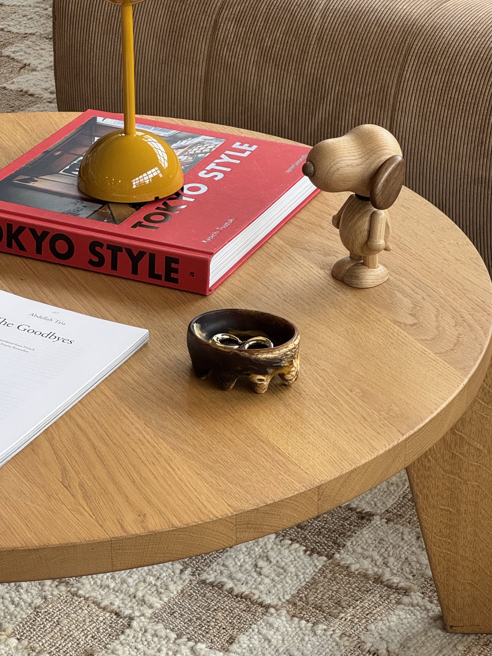 A wooden coffee table displays a TOKYO STYLE book, yellow lamp, wooden dog figurine, white magazine, and the Dinosaur Designs Crown Dish by Dinosaur Designs holding rings for a sculptural accent atop the patterned carpet.