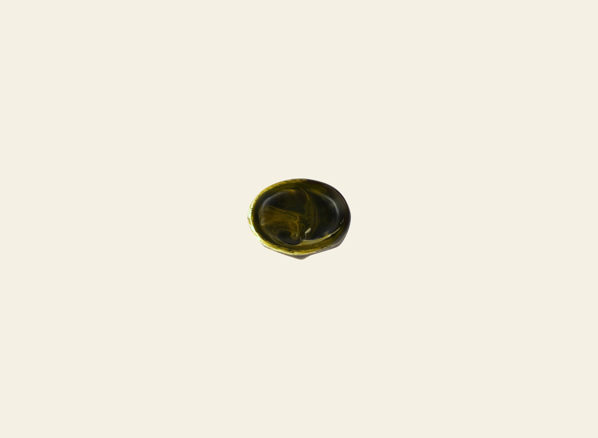 A small, circular swirl of dark green liquid rests at the center of a plain light cream surface, echoing the sculptural elegance of the Dinosaur Designs Crown Dish by Dinosaur Designs.