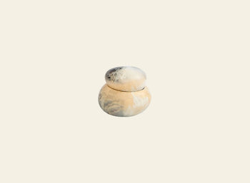The Dinosaur Designs Small Pebble Jar features a matching lid and round, sculptural design with smooth swirls of beige, cream, and light gray tones, displayed against a plain off-white background.