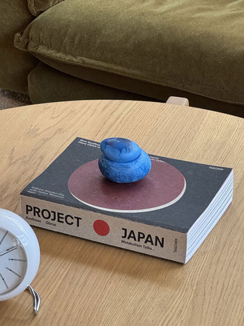 A blue rubber duck rests atop a thick book titled PROJECT JAPAN on a wooden table. Nearby, the Dinosaur Designs Small Pebble Jar by Dinosaur Designs brings stylish storage, with an analog clock and green couch partially visible in the background.