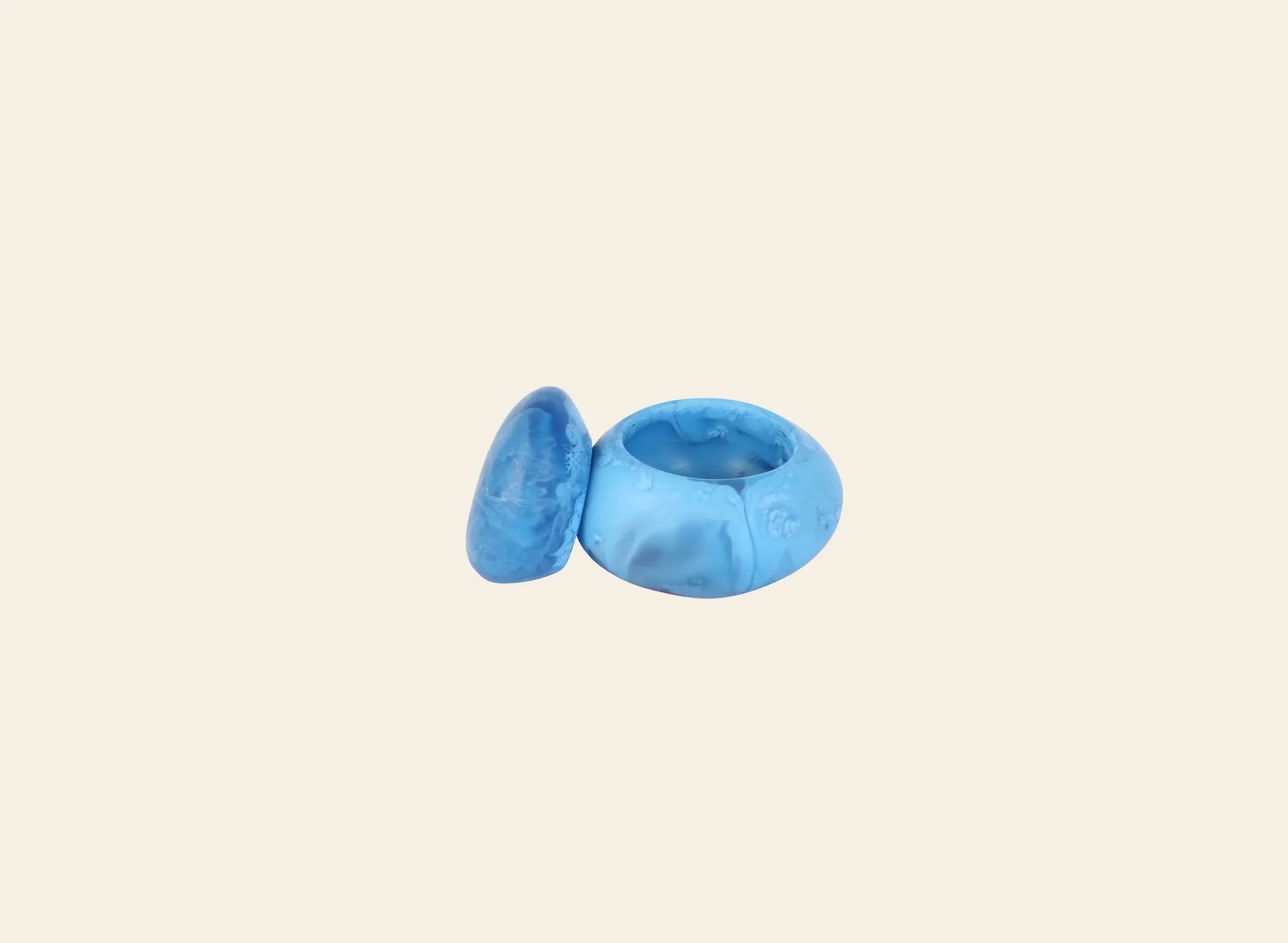 A chunky blue resin ring with an uneven, organic shape is displayed alongside the Dinosaur Designs Small Pebble Jar, a matching sculptural accessory for stylish storage, both set against a plain light beige background.