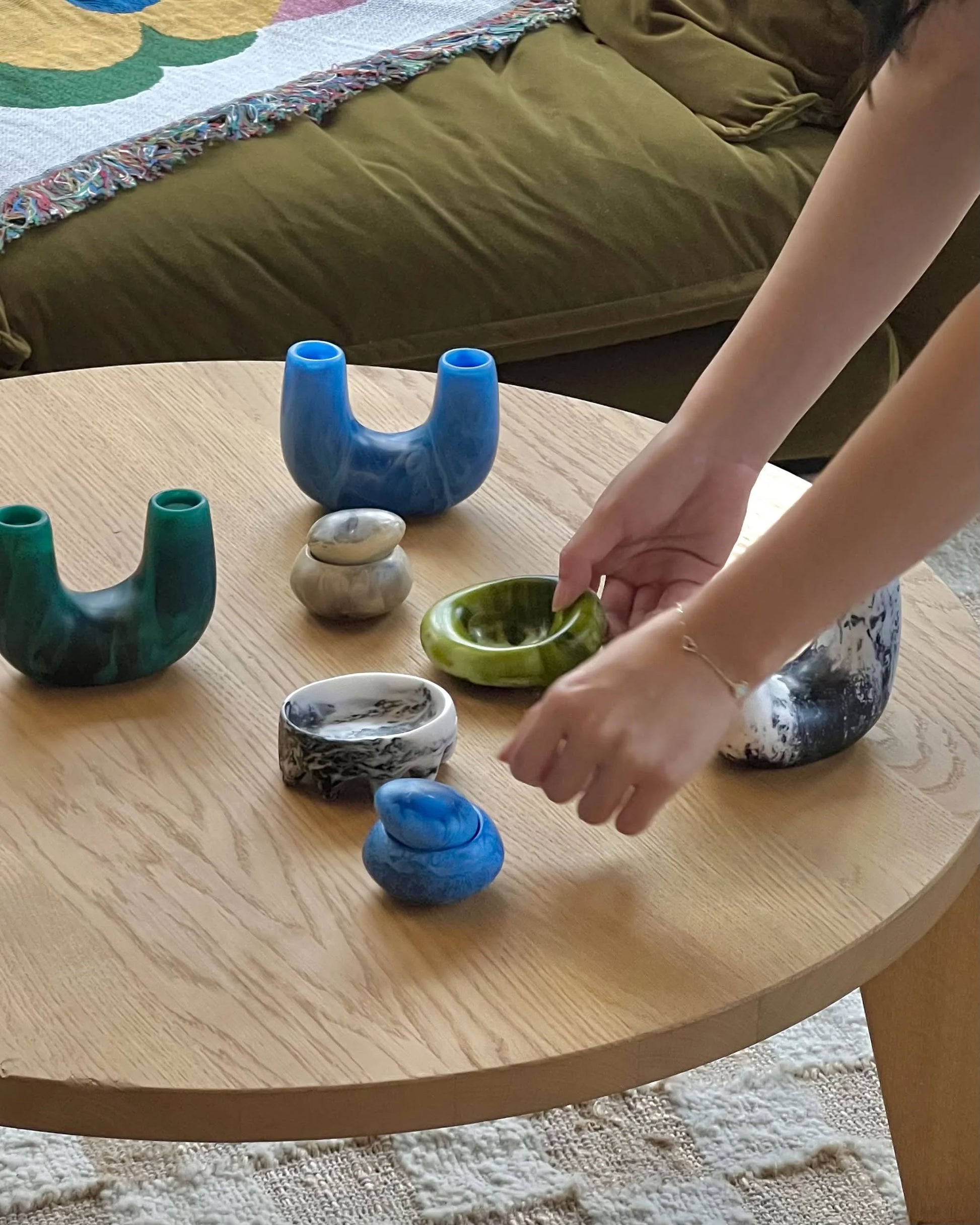 A person arranges colorful, uniquely shaped ceramic and stone objects, including the Dinosaur Designs Small Pebble Jar, on a round wooden coffee table. A green sofa with a patterned blanket appears in the background.