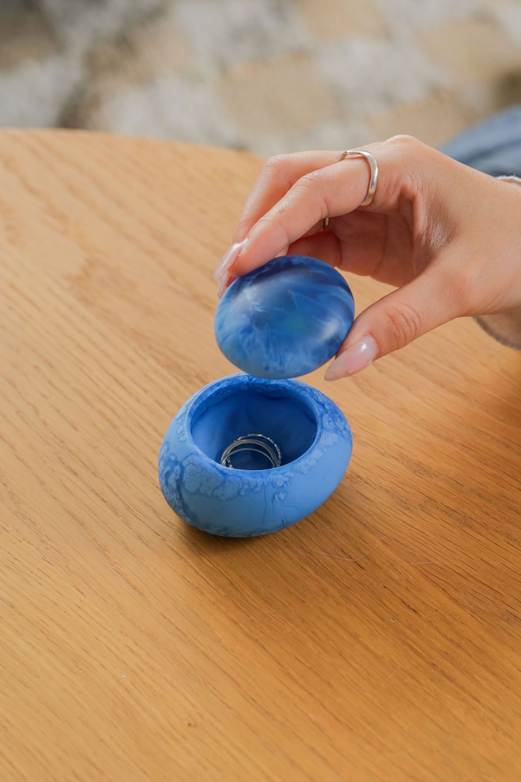 A hand with a silver ring lifts the lid off a Dinosaur Designs Small Pebble Jar, featuring a round blue marbled design on a wooden surface, revealing another silver ring stylishly stored inside.