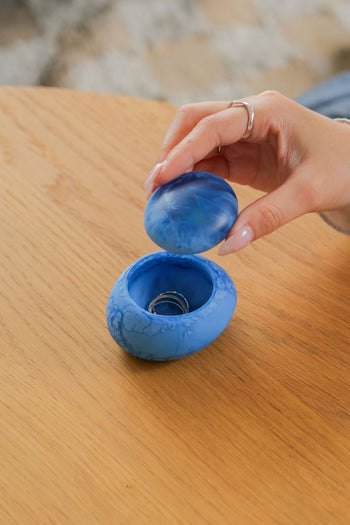 A hand with a silver ring lifts the lid off a Dinosaur Designs Small Pebble Jar, featuring a round blue marbled design on a wooden surface, revealing another silver ring stylishly stored inside.