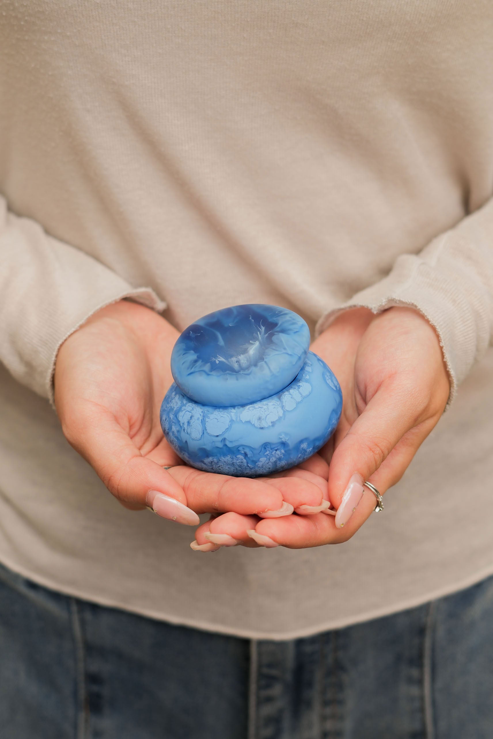 A person in a light beige long-sleeve shirt and jeans holds the Dinosaur Designs Small Pebble Jar—a round, blue, marbled storage piece with a puckered center lid from Dinosaur Designs—in both hands.