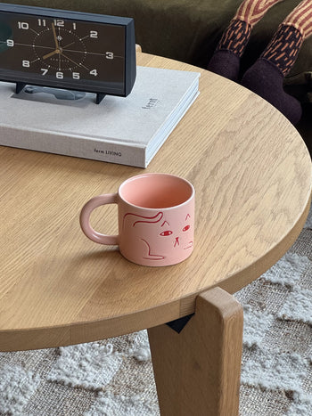 The Donna Wilson Cat Mug by Donna Wilson, featuring a minimalist pink cat face design, rests on a wooden coffee table beside a book and clock, with cozy socked feet in the background—ideal for relaxing coffee moments.