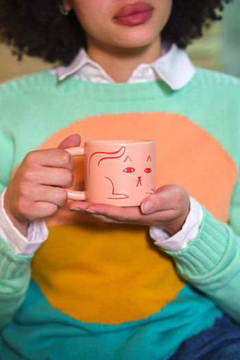 Someone enjoys a coffee break, holding a Donna Wilson Cat Mug—a pink mug with a red cartoon face—while wearing a green, orange, and blue sweater. Only the person’s hands and torso are visible; the mug is by Donna Wilson.