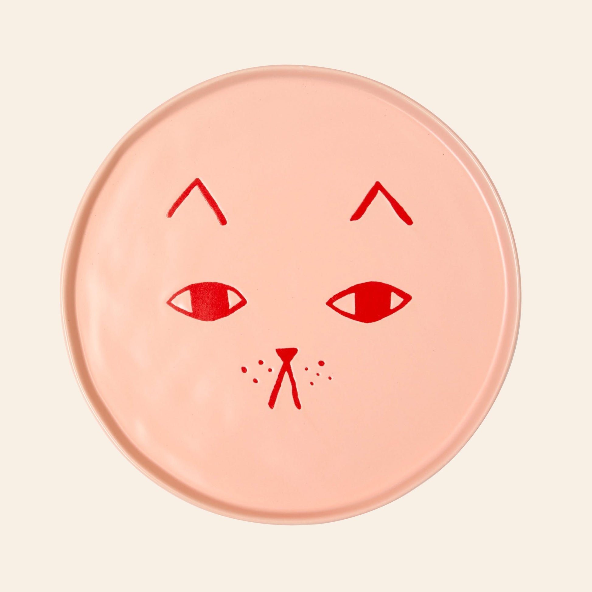 The Donna Wilson Cat Plate by Donna Wilson is a round, light pink ceramic plate with a minimalist red cat face—simple eyes, nose, mouth, whiskers, and eyebrows—ideal as charming cat-themed decor.