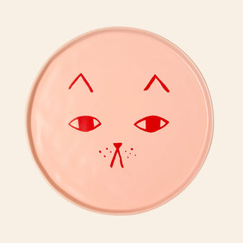The Donna Wilson Cat Plate by Donna Wilson is a round, light pink ceramic plate with a minimalist red cat face—simple eyes, nose, mouth, whiskers, and eyebrows—ideal as charming cat-themed decor.