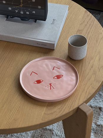 A round wooden table with a Donna Wilson Cat Plate featuring a cat face, a small ceramic cup, and a rectangular book topped with a clock.