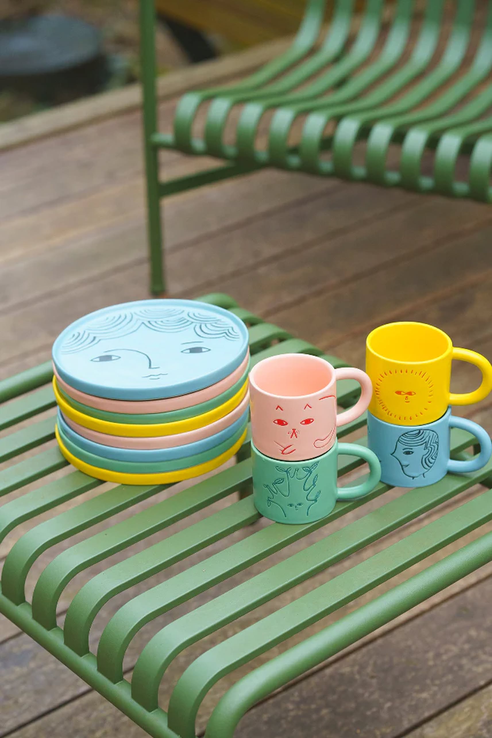 Four colorful plates and four mugs, including the Donna Wilson Cat Plate by Donna Wilson with whimsical faces, are stacked on a green slatted outdoor bench with a matching chair in the background on a wooden deck.