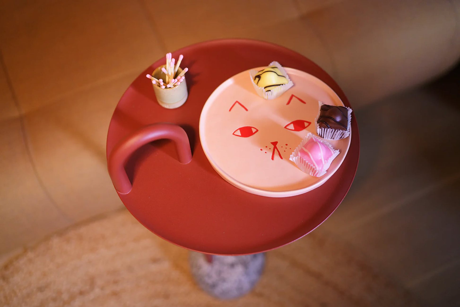 A round red side table displays a Donna Wilson Cat Plate by Donna Wilson, featuring a simple cat face and three pastries, beside a small cup of toothpicks, set against a blurred indoor background.