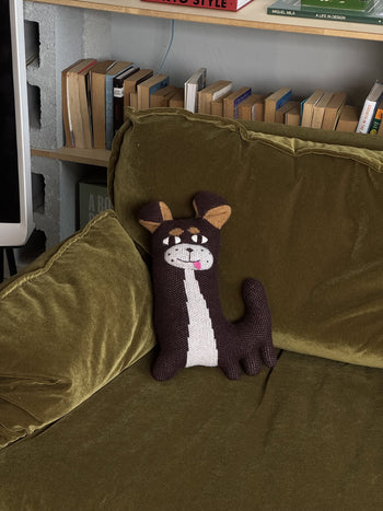 A Donna Wilson Creature - Chip Chihuahua plushie sits on an olive green velvet couch, with books on shelves visible in the background.