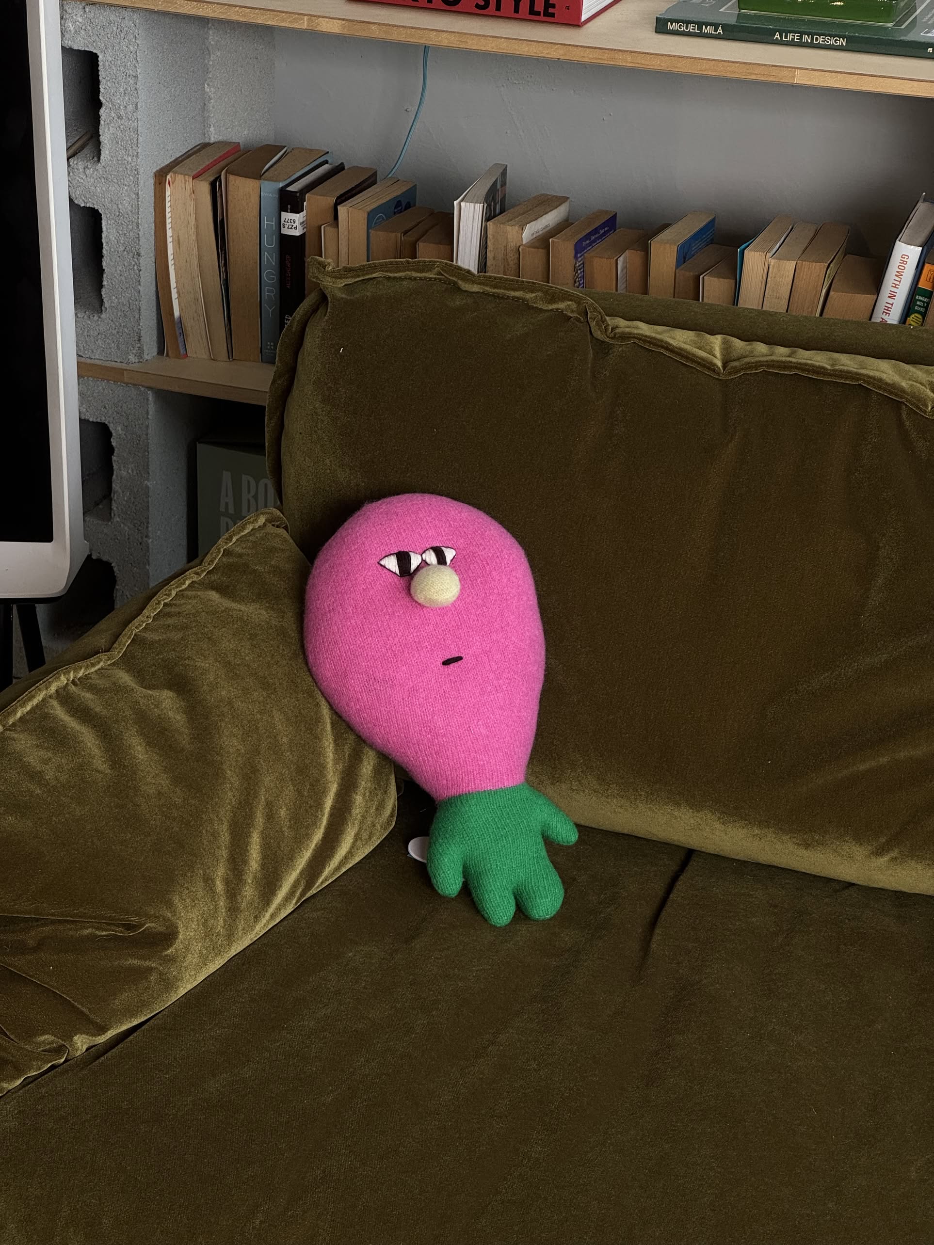 A Donna Wilson Creature - Claude plush from Donna Wilson, in pink and green with a face, leans against an olive-green velvet couch; books are arranged on a shelf in the background.