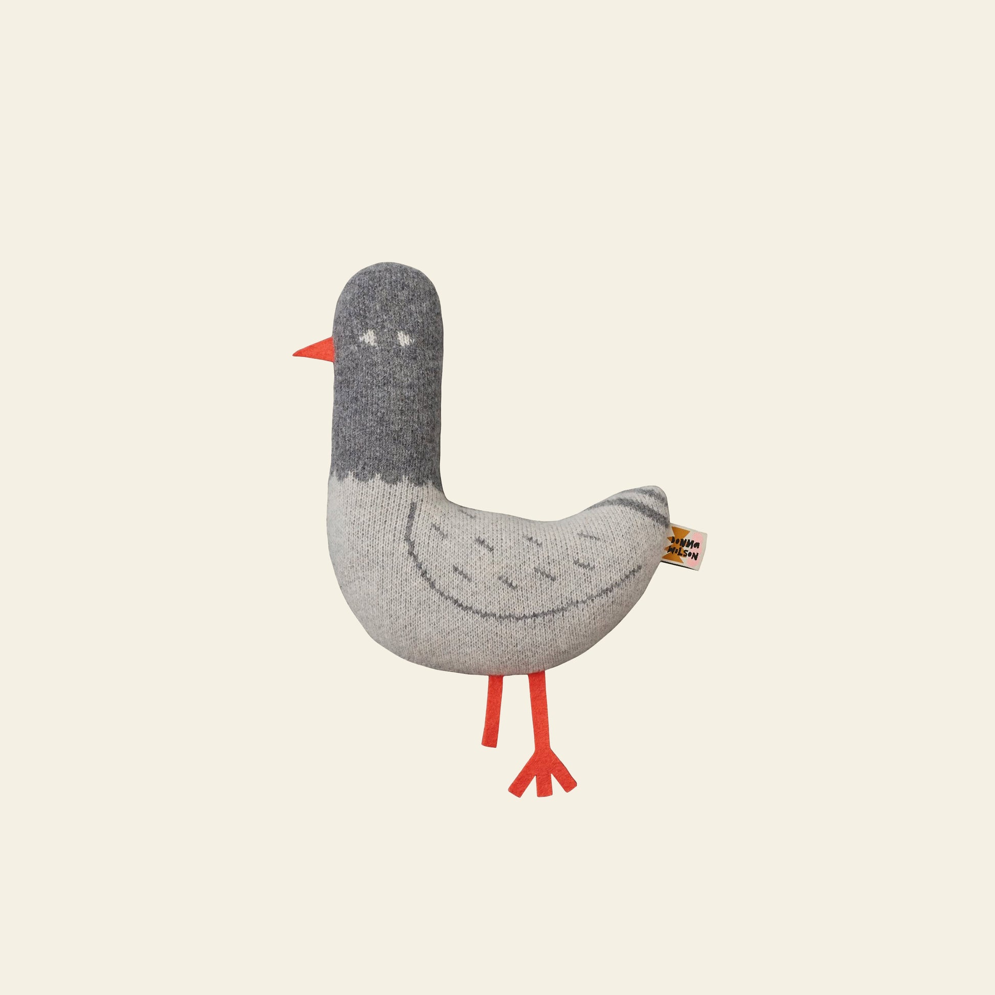 Meet the Donna Wilson Creature - Paddy Pigeon, a hand-assembled plush toy by Donna Wilson. This lambswool pigeon has a gray body, darker gray head, orange beak and legs, and stands upright on a light background.