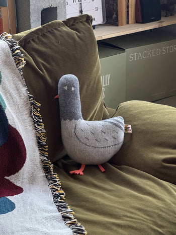 A hand-assembled Donna Wilson Creature - Paddy Pigeon plush toy with pink feet sits on an olive green couch beside a fringed multicolored blanket, with boxes and books in the background.
