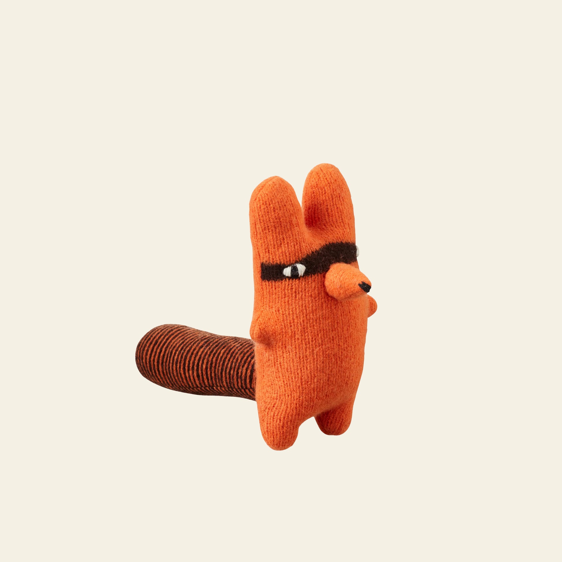 The Donna Wilson Creature - Rill is a small, orange lambswool plush from Donna Wilson shaped like an animal with round ears, a brown mask around its eyes, and a striped tail, shown upright against a plain light background.
