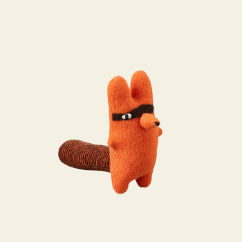 The Donna Wilson Creature - Rill is a small, orange lambswool plush from Donna Wilson shaped like an animal with round ears, a brown mask around its eyes, and a striped tail, shown upright against a plain light background.