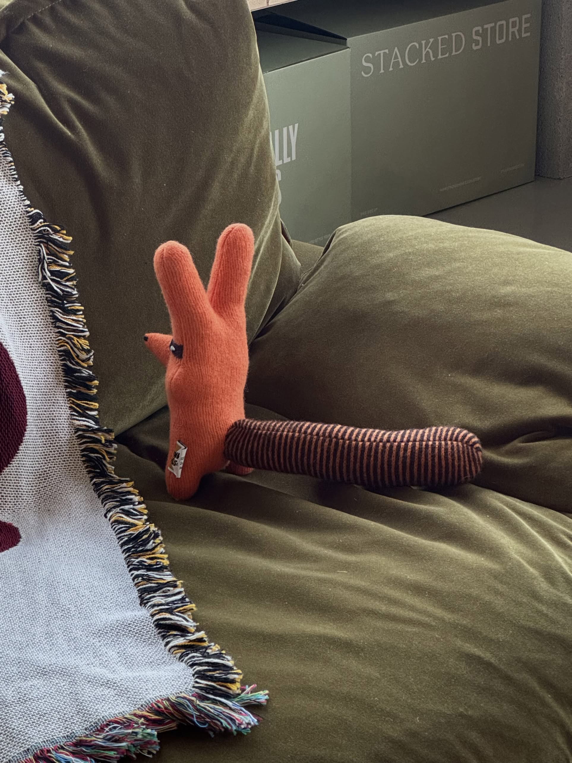 The Donna Wilson Creature - Rill, a soft lambswool toy resembling an orange animal with long ears and a striped brown tail, rests on an olive green couch beside a fringed white blanket. Cardboard boxes are stacked in the background.