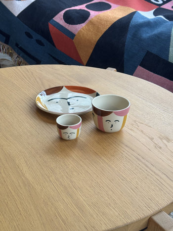 A Donna Wilson Freya Face Cup, a medium artisanal coffee cup, and a plate with abstract face designs sit on a light wooden table, with colorful geometric-patterned fabric in the background.