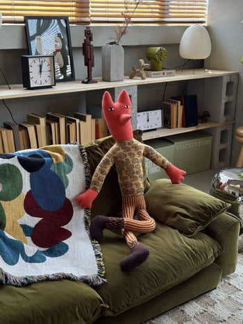 The Donna Wilson Mini Giant Creature - Orange Fox, handcrafted by Donna Wilson, wears a patterned shirt and striped pants while sitting on a green velvet couch in a cozy, colorful living room filled with books and decor.