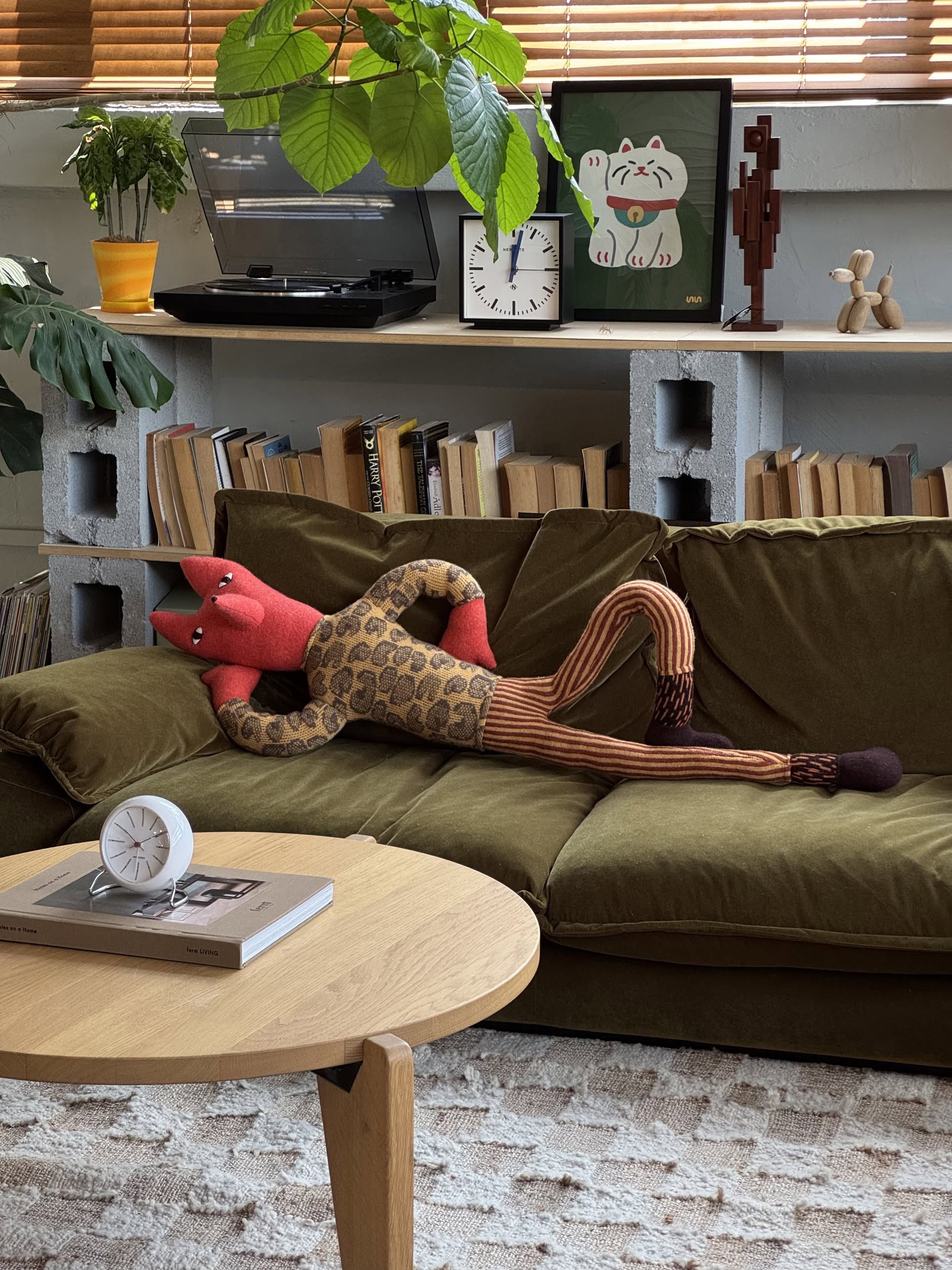 The Donna Wilson Mini Giant Creature - Orange Fox plush lounges on a green velvet sofa in a cozy living room filled with books, plants, and playful decor on the shelves behind.