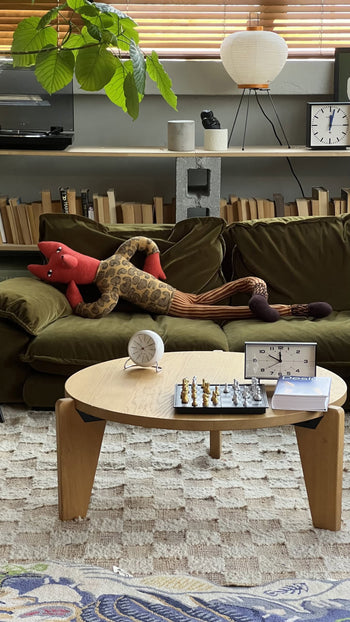 A cozy living room with a green sofa featuring the Donna Wilson Mini Giant Creature - Orange Fox, a sustainable plush toy, plus a round wooden coffee table with chess set and clock, books on shelves, and houseplants by a window with wooden blinds.