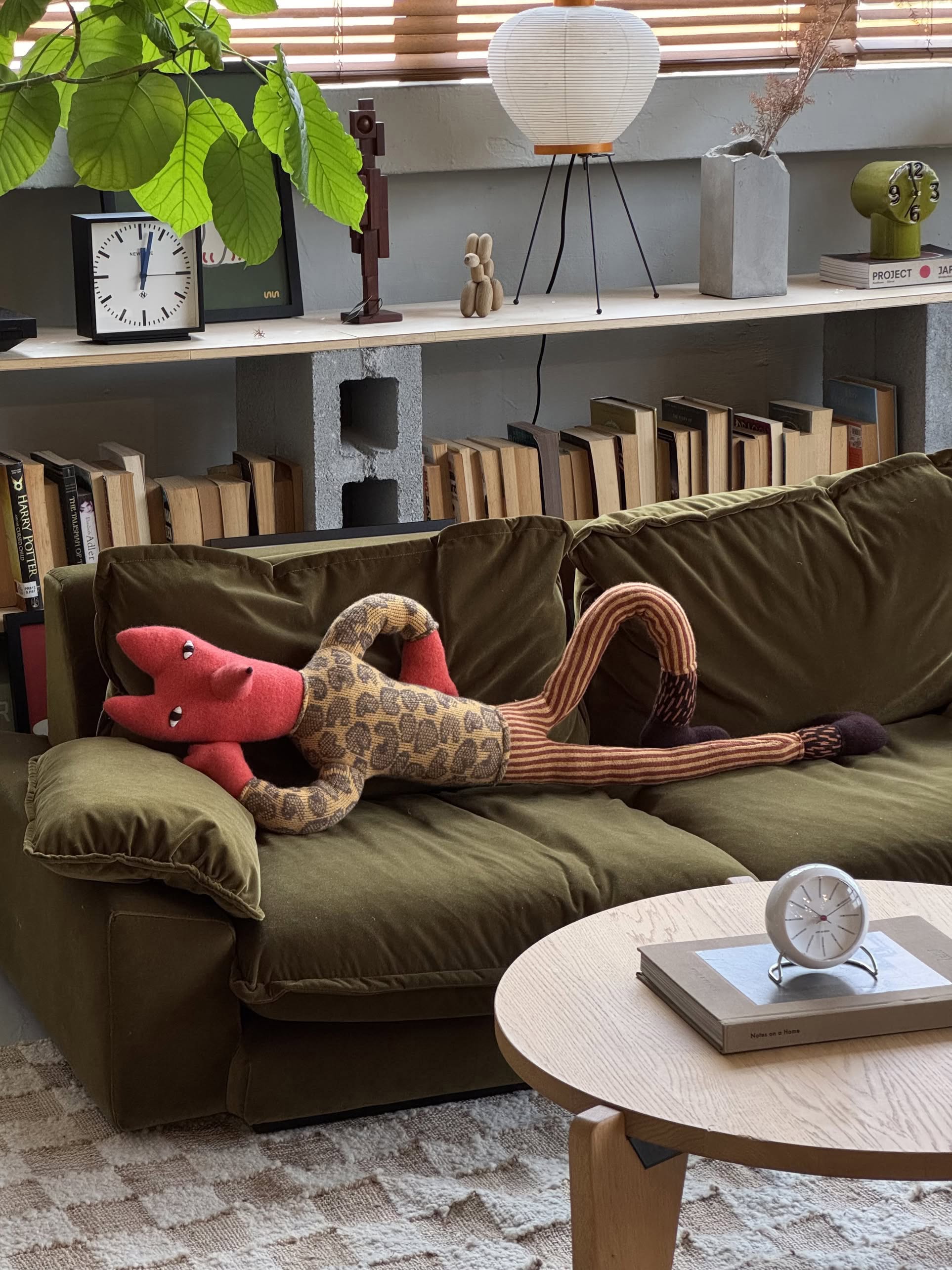 The Donna Wilson Mini Giant Creature - Orange Fox, a handcrafted plush toy, lounges on a green sofa in a cozy living room with books, a clock, and potted plants displayed on shelves in the background.