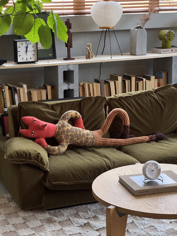 The Donna Wilson Mini Giant Creature - Orange Fox, a handcrafted plush toy, lounges on a green sofa in a cozy living room with books, a clock, and potted plants displayed on shelves in the background.