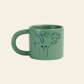 The Donna Wilson Tree Mug by Donna Wilson features a whimsical, line-drawn tree with a face and finger-like branches debossed on moss-green ceramic, creating a striking contrast against its off-white background.